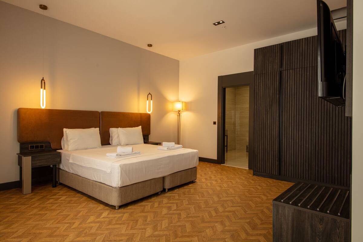 A hotel room with a bed, bedside tables, a TV, and a wooden floor.