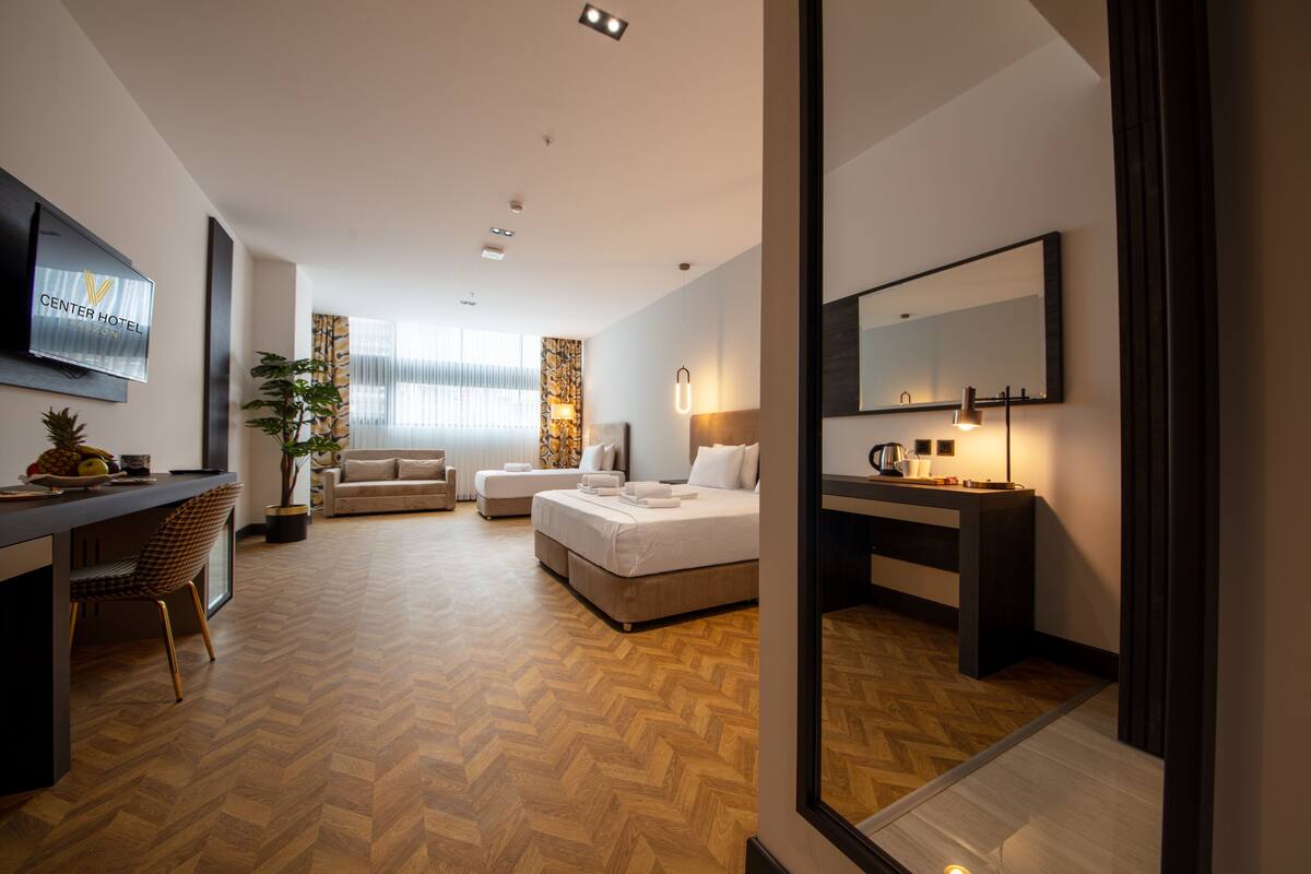 A modern hotel room with a bed, a desk, a sofa, a TV, and a large mirror.