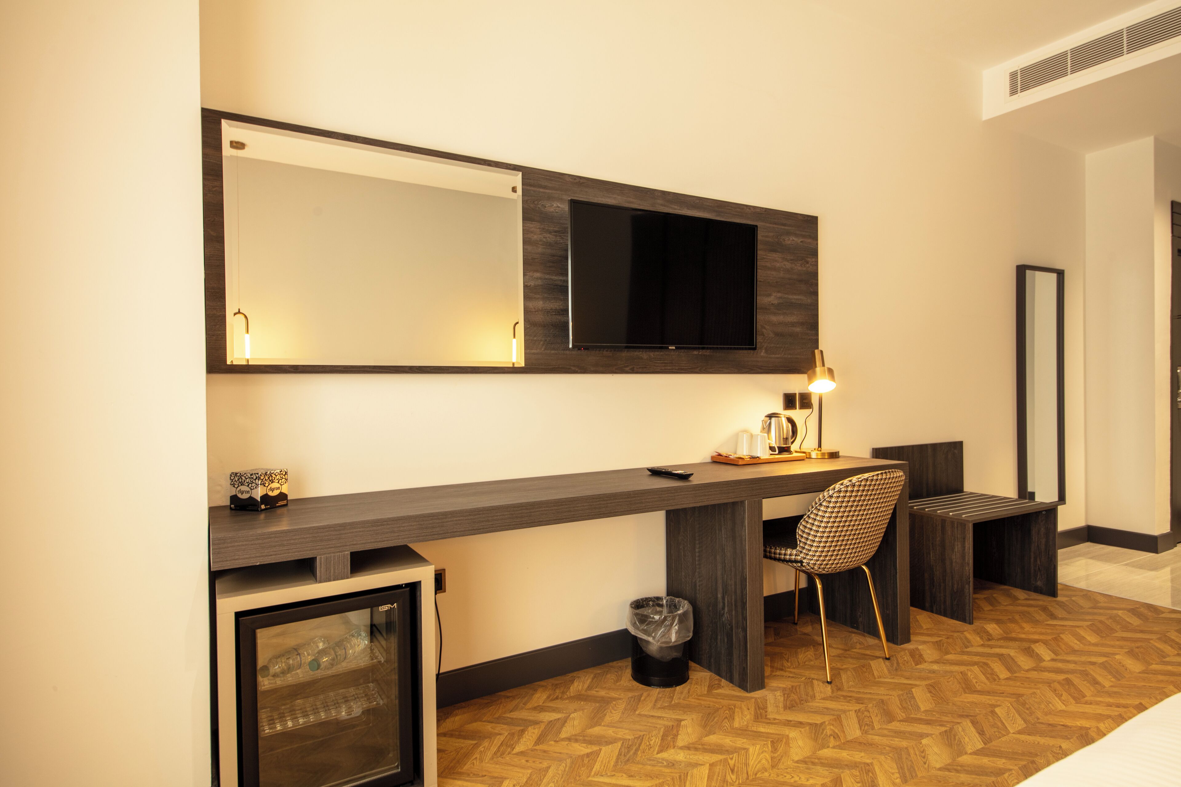 Habitación de hotel con un escritorio grande, una silla, un televisor montado en la pared, un refrigerador pequeño y un cesto de basura.