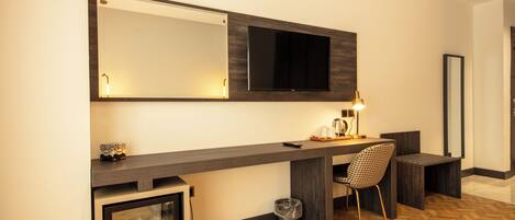 Habitación de hotel con un escritorio grande, una silla, un televisor montado en la pared, un refrigerador pequeño y un cesto de basura.