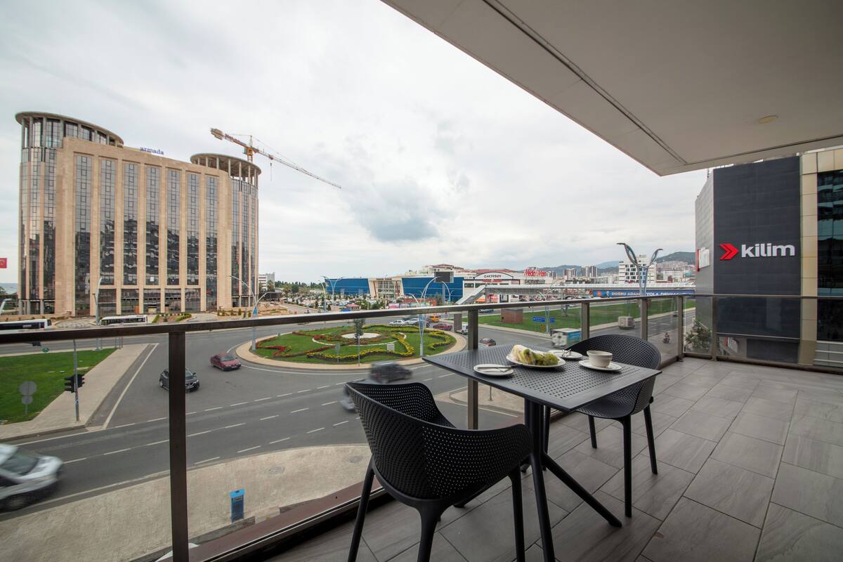 A balcony with a table and chairs, overlooking a cityscape with buildings and a roundabout.