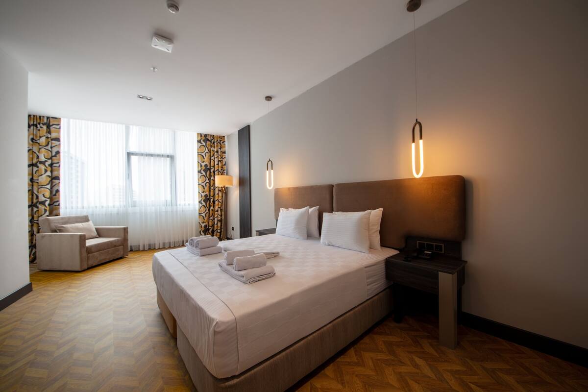 A hotel room with a large bed, a bedside table, a chair, and a window with curtains.