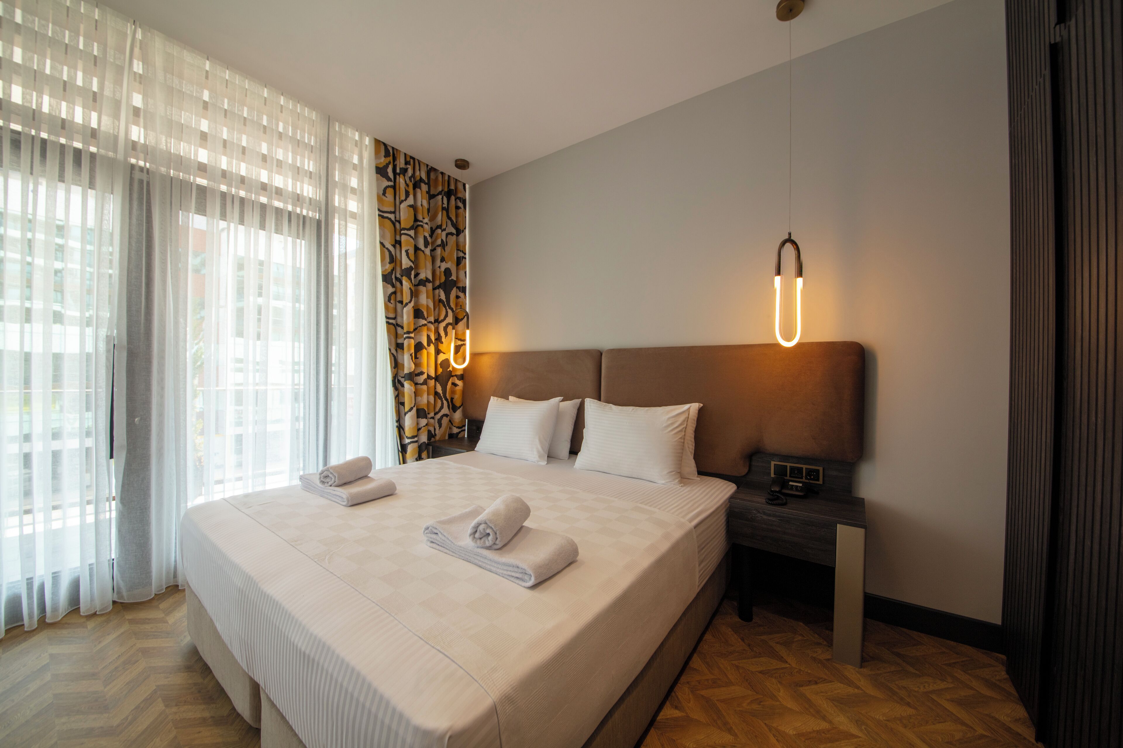 Quarto de hotel com uma cama grande, uma mesa de cabeceira e um candeeiro fixo na parede.