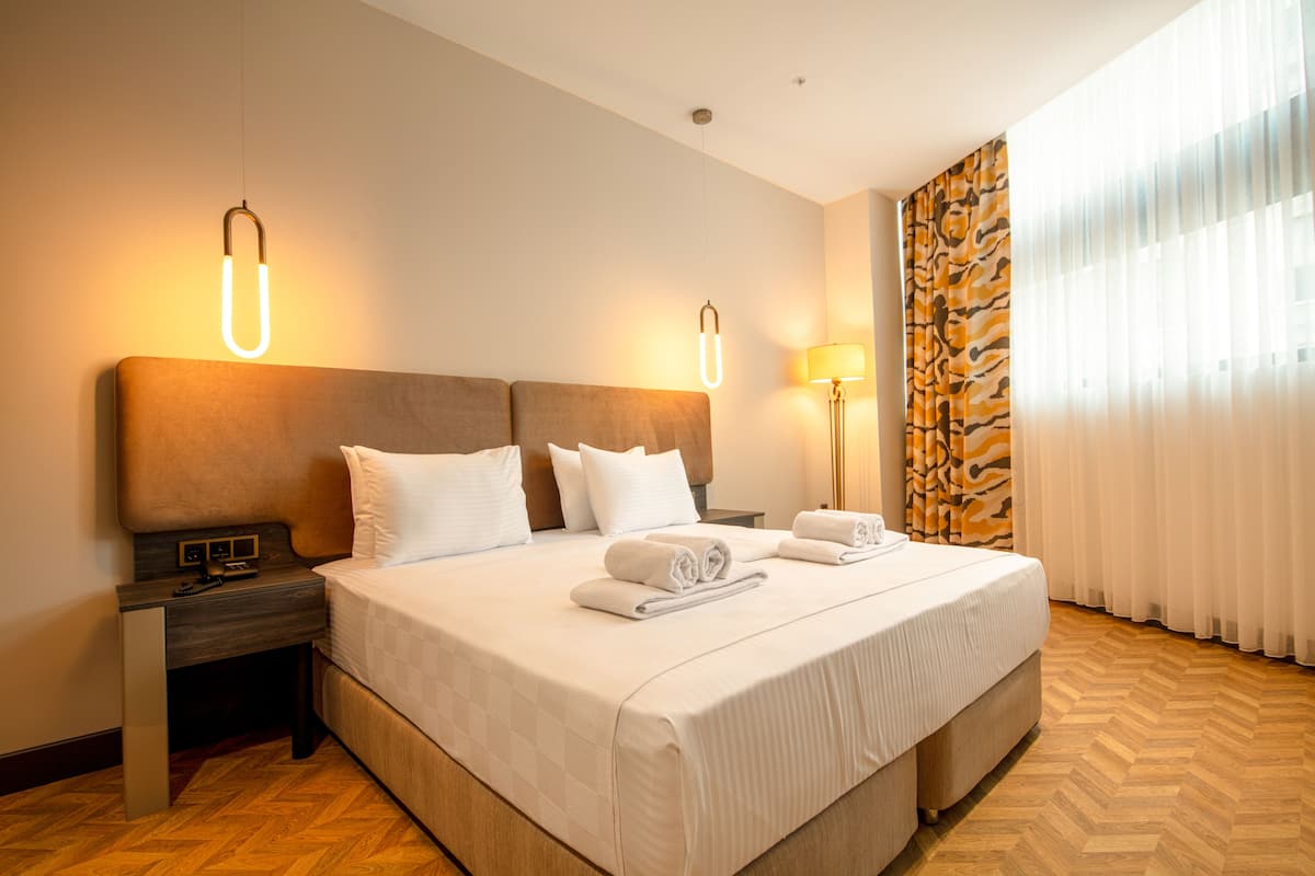 A hotel room with a large bed, two bedside tables, a nightstand, a lamp, and a window with curtains.