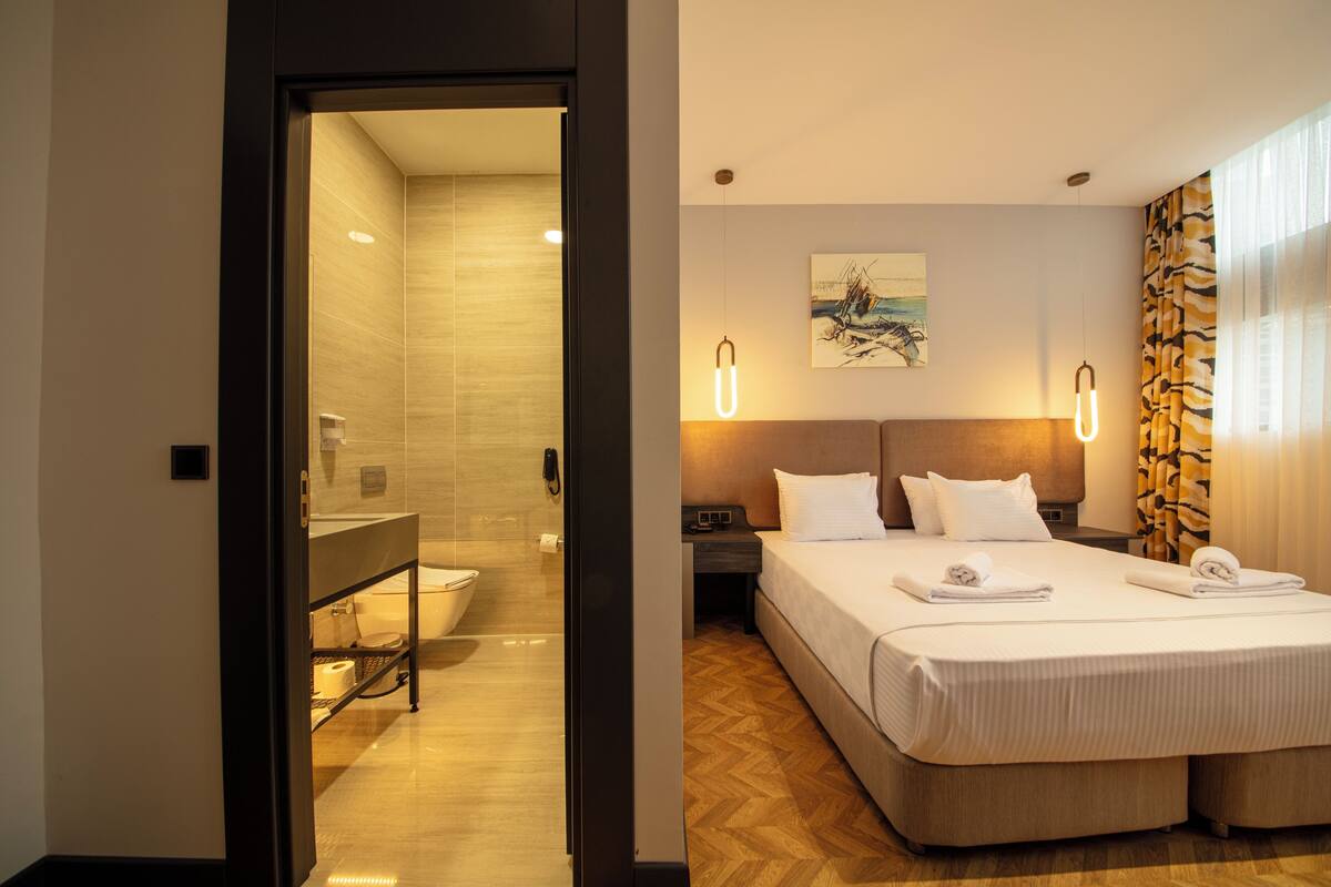 A hotel room with a wooden floor, a bed with white linens, a nightstand, a wall-mounted light fixture, and a painting above the bed.