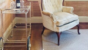 Een kamer met een antieke fauteuil, een glazen karretje met wijnglazen en een fles, en een raam met luiken.