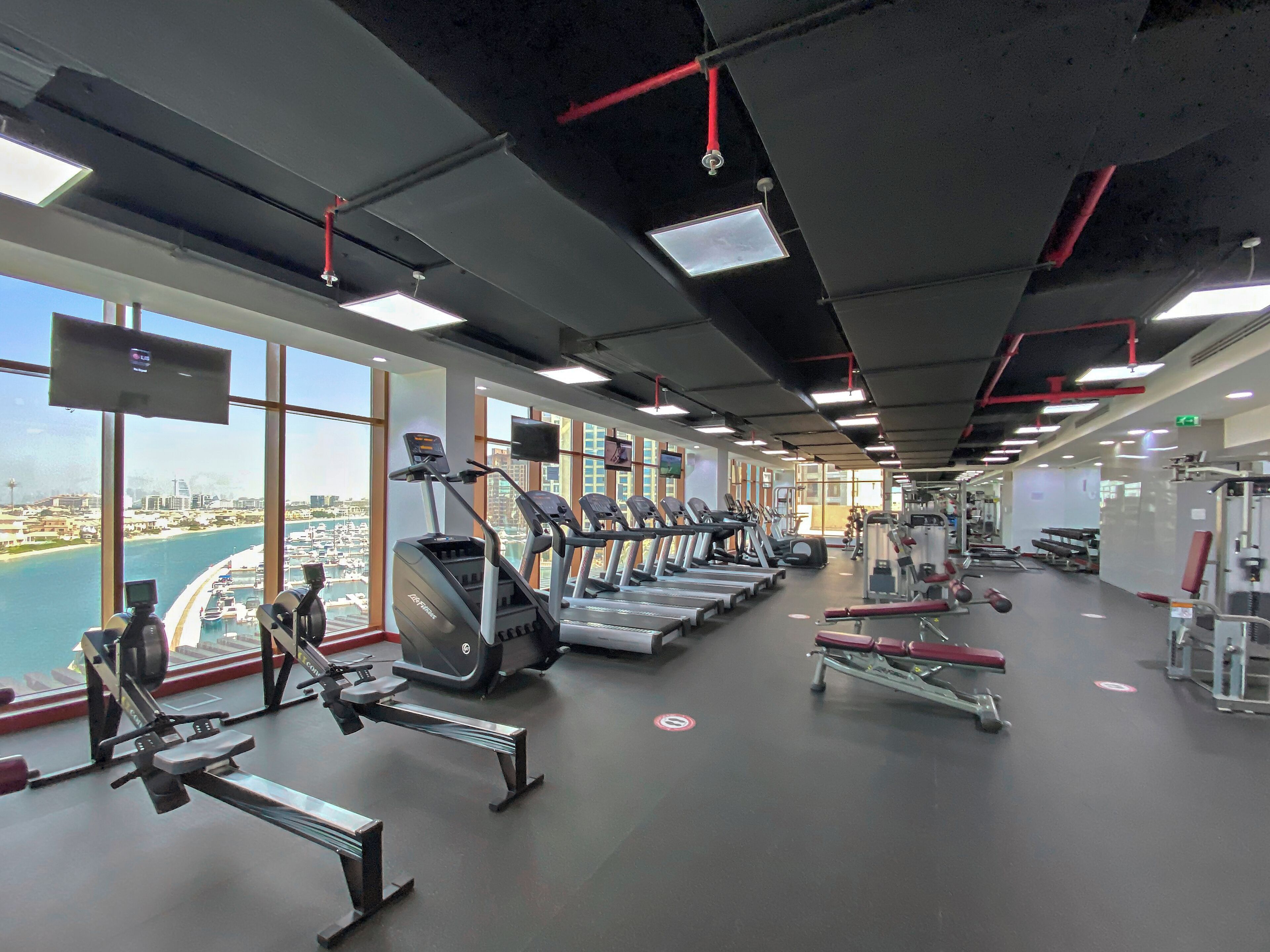 A gym with treadmills, weight machines, and a view of a cityscape through large windows.