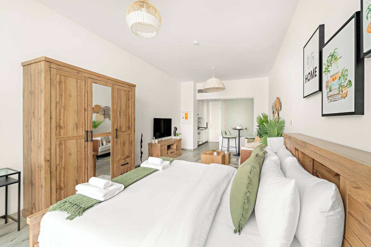 A modern hotel room with a large bed, wooden wardrobe, a television, a desk, and a dining area.