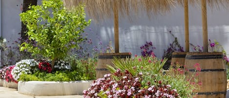 A garden with various potted plants and flowers, a wooden barrel, and a thatched roof structure.
