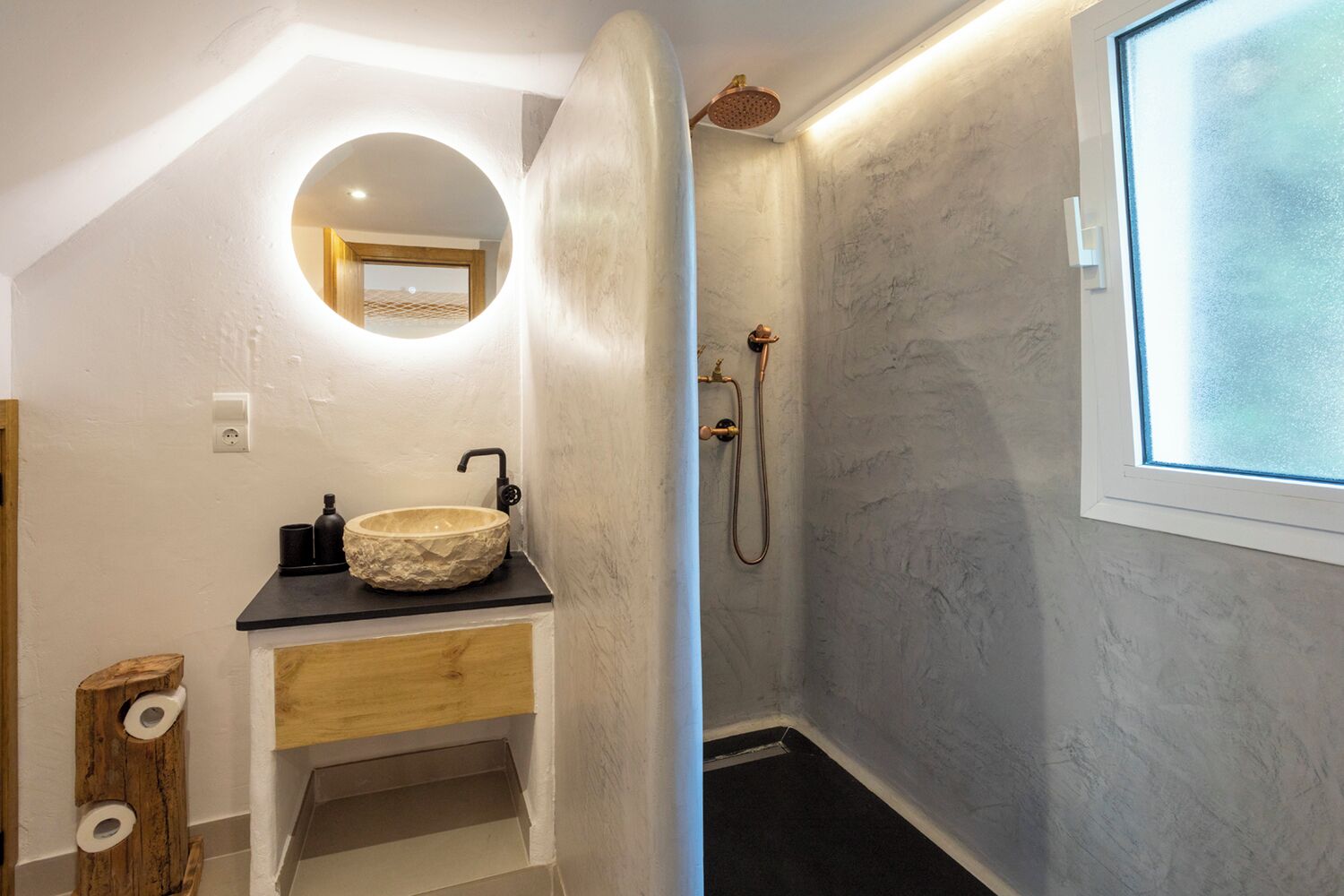 A bathroom with a round mirror, a wooden vanity, a stone sink, a shower with a handheld showerhead, and a window with frosted glass.