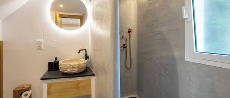 A bathroom with a round mirror, a wooden vanity, a stone sink, a shower with a handheld showerhead, and a window with frosted glass.