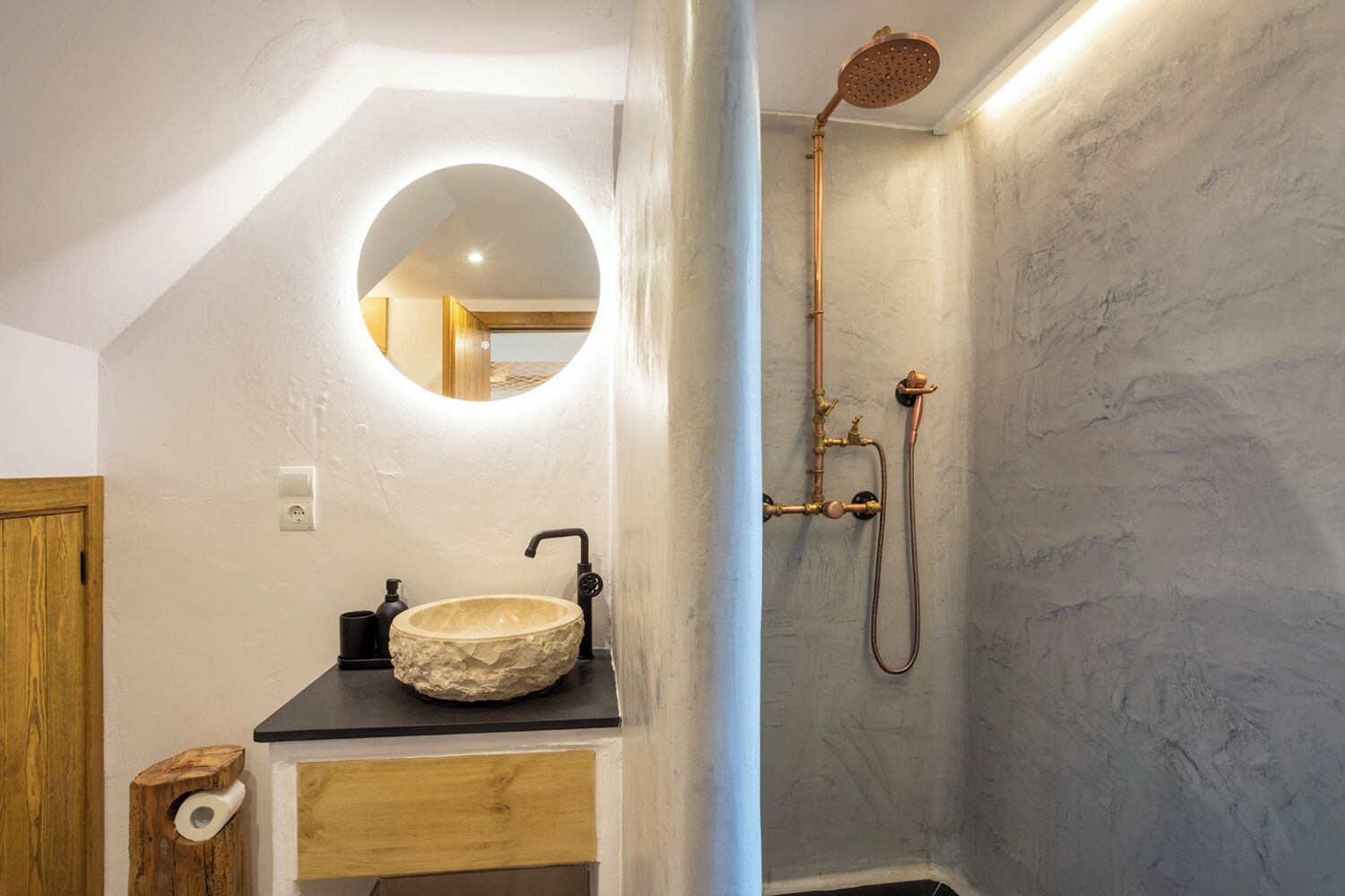 Une salle de bain avec un miroir rond, un lavabo en pierre et une douche équipée de robinetterie en cuivre.