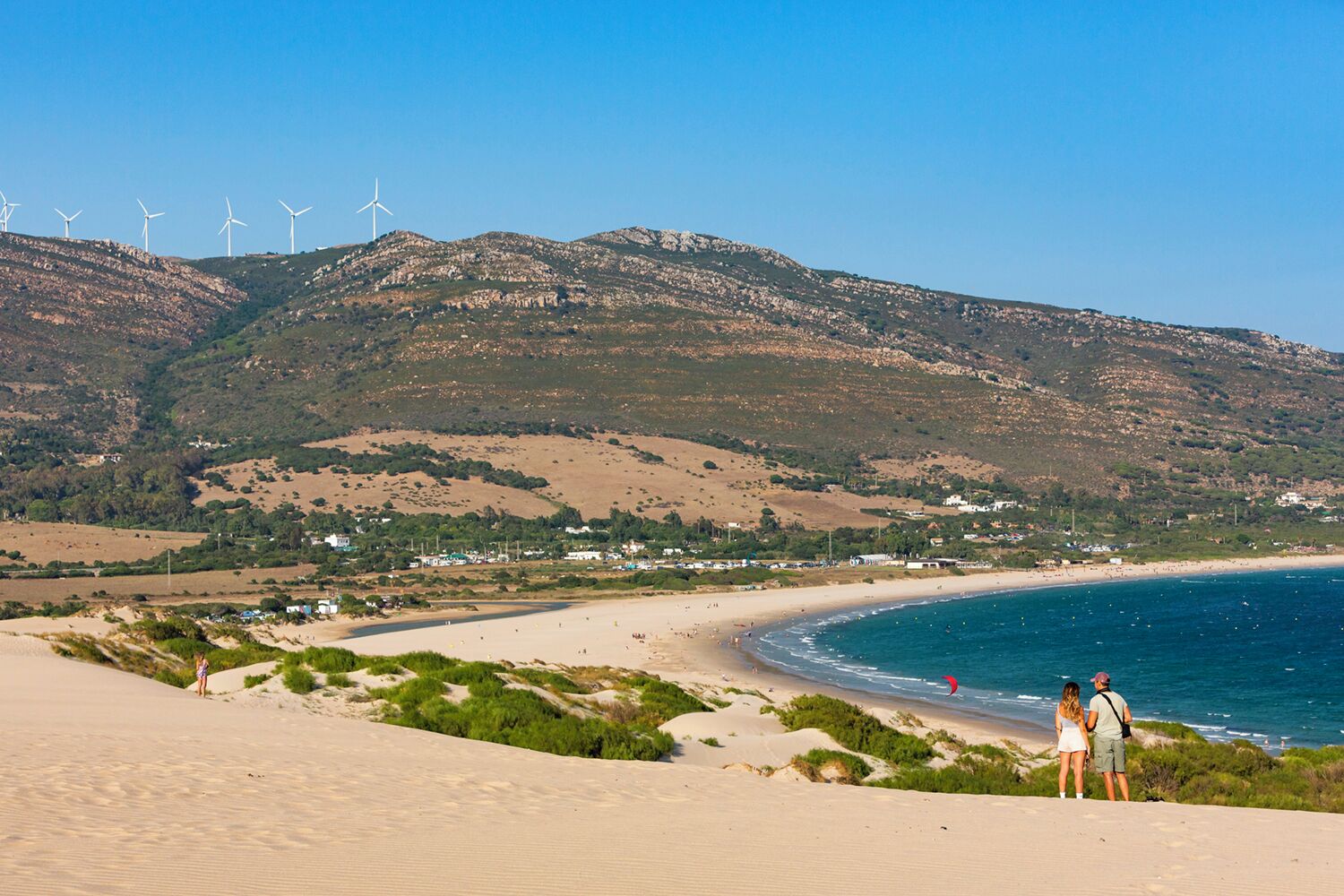 Ein Strand mit Dünen, blauem Meer und Windkraftanlagen im Hintergrund.