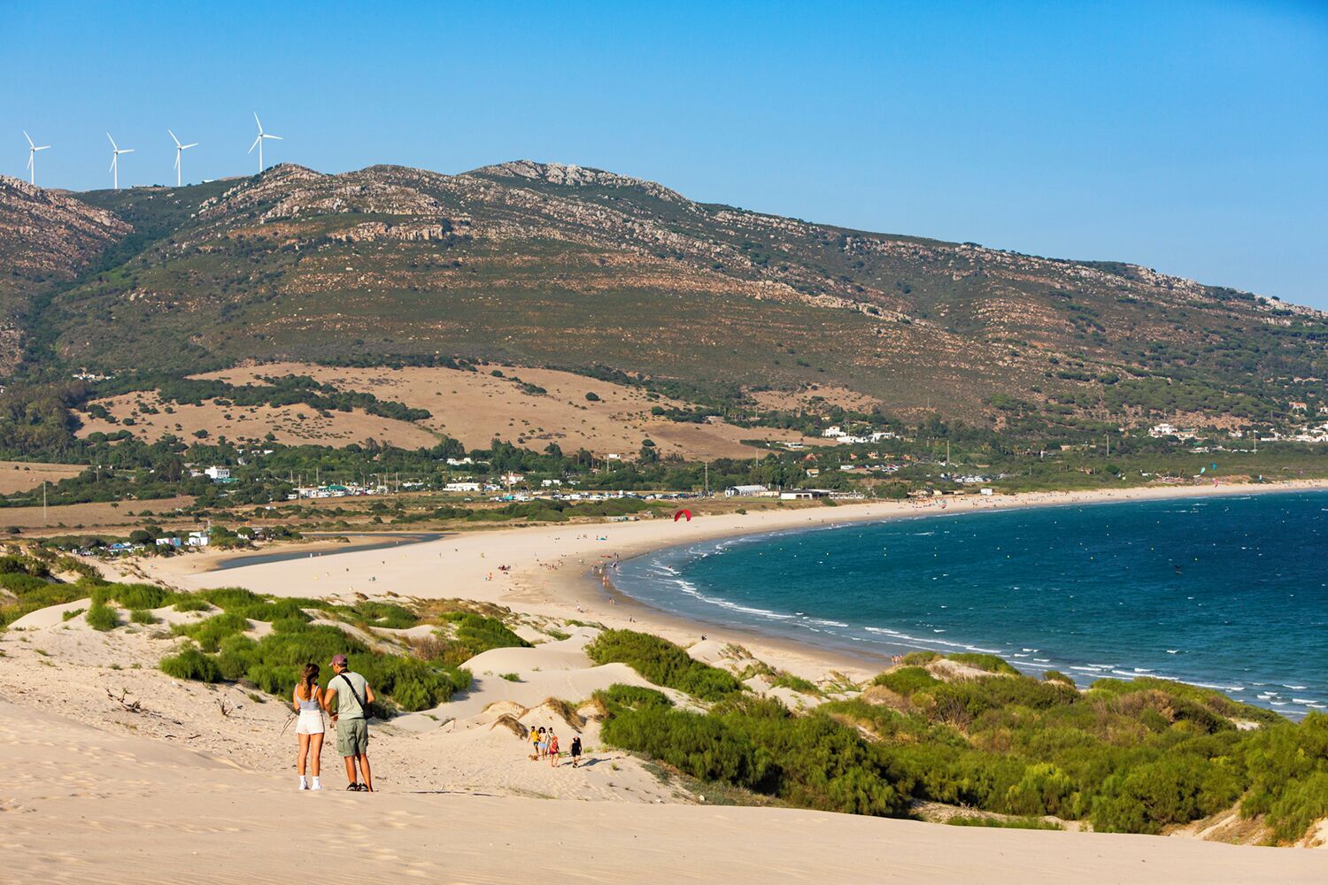 Un paisaje costero con una playa arenosa, un mar azul cristalino y aerogeneradores al fondo.