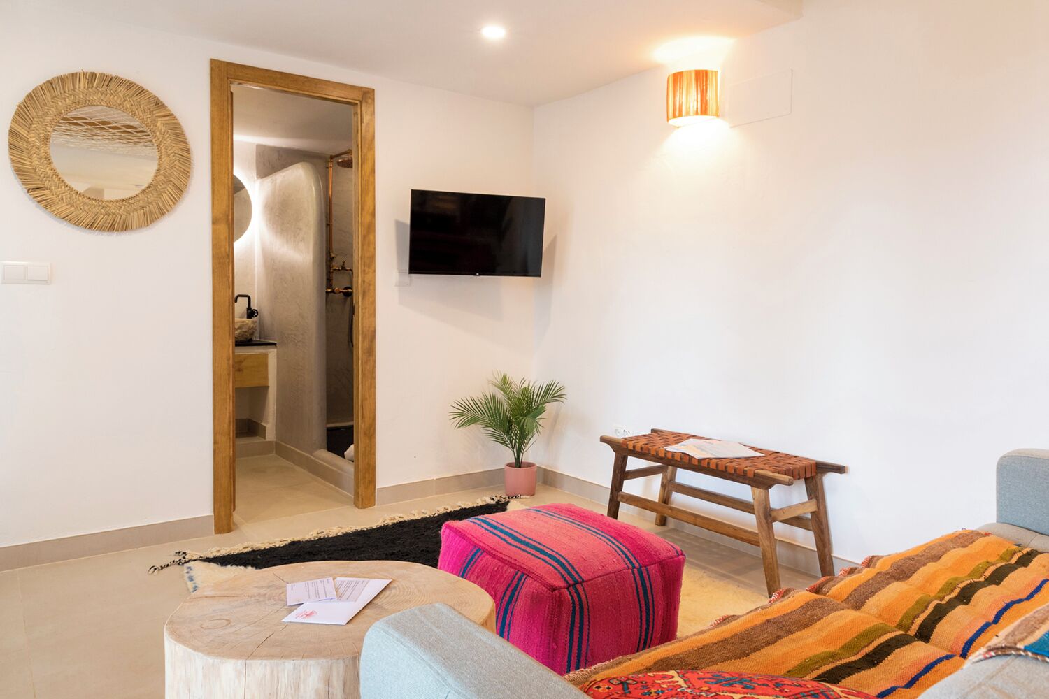Un salón moderno con un banco de madera, un televisor montado en la pared, un espejo redondo y una planta en maceta.