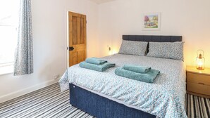 A bedroom with a bed, a wooden door, a nightstand, a lamp, and a window with curtains. - Sole Bay Cottage (Lowestoft)
