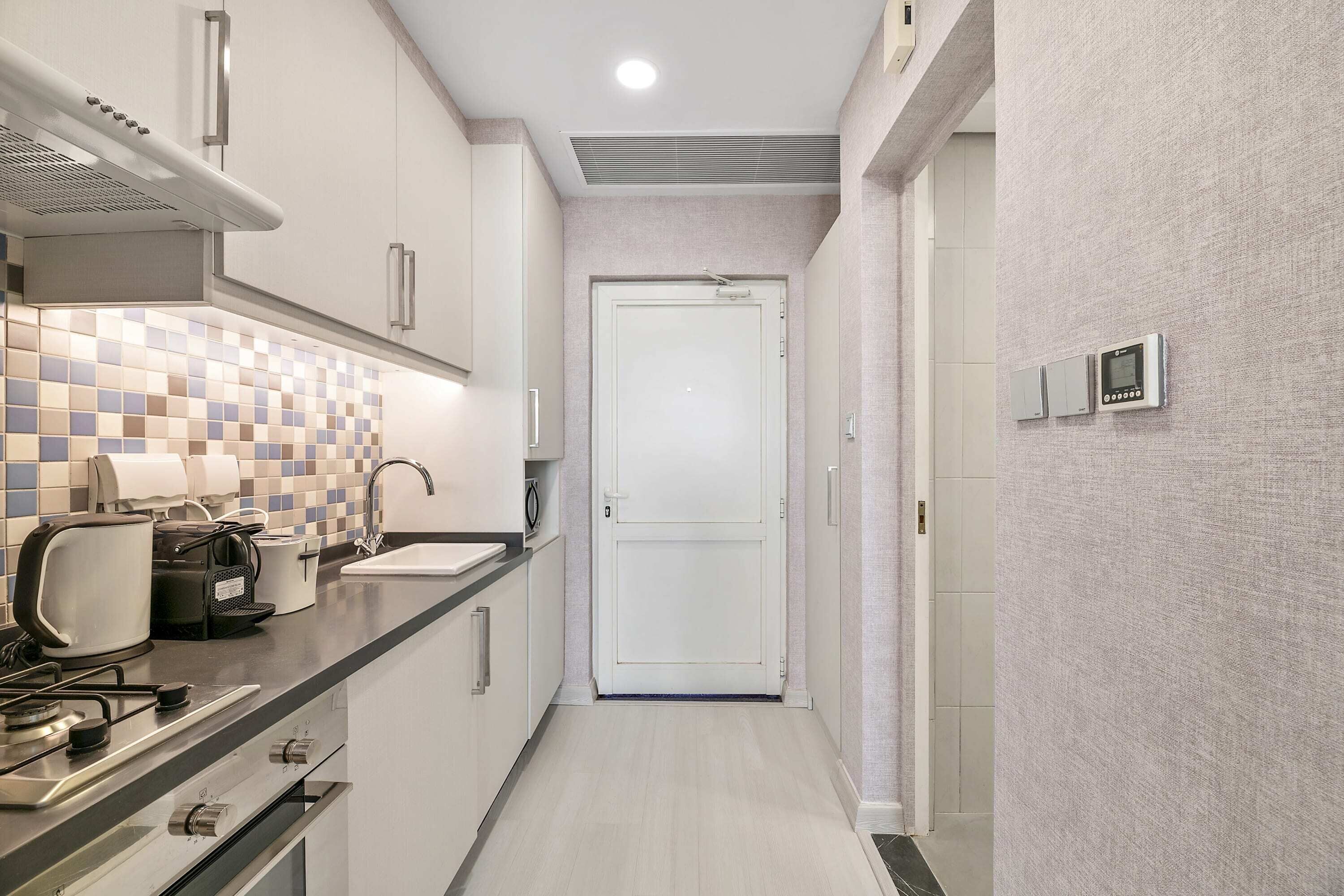 A narrow kitchen with a tiled backsplash, a stainless steel range hood, and a white door at the end.