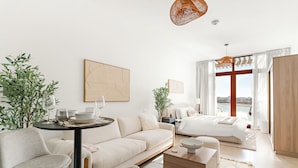 Une chambre d’hôtel moderne avec un canapé blanc, une table à manger, un lit donnant sur une vue agréable et une grande fenêtre.