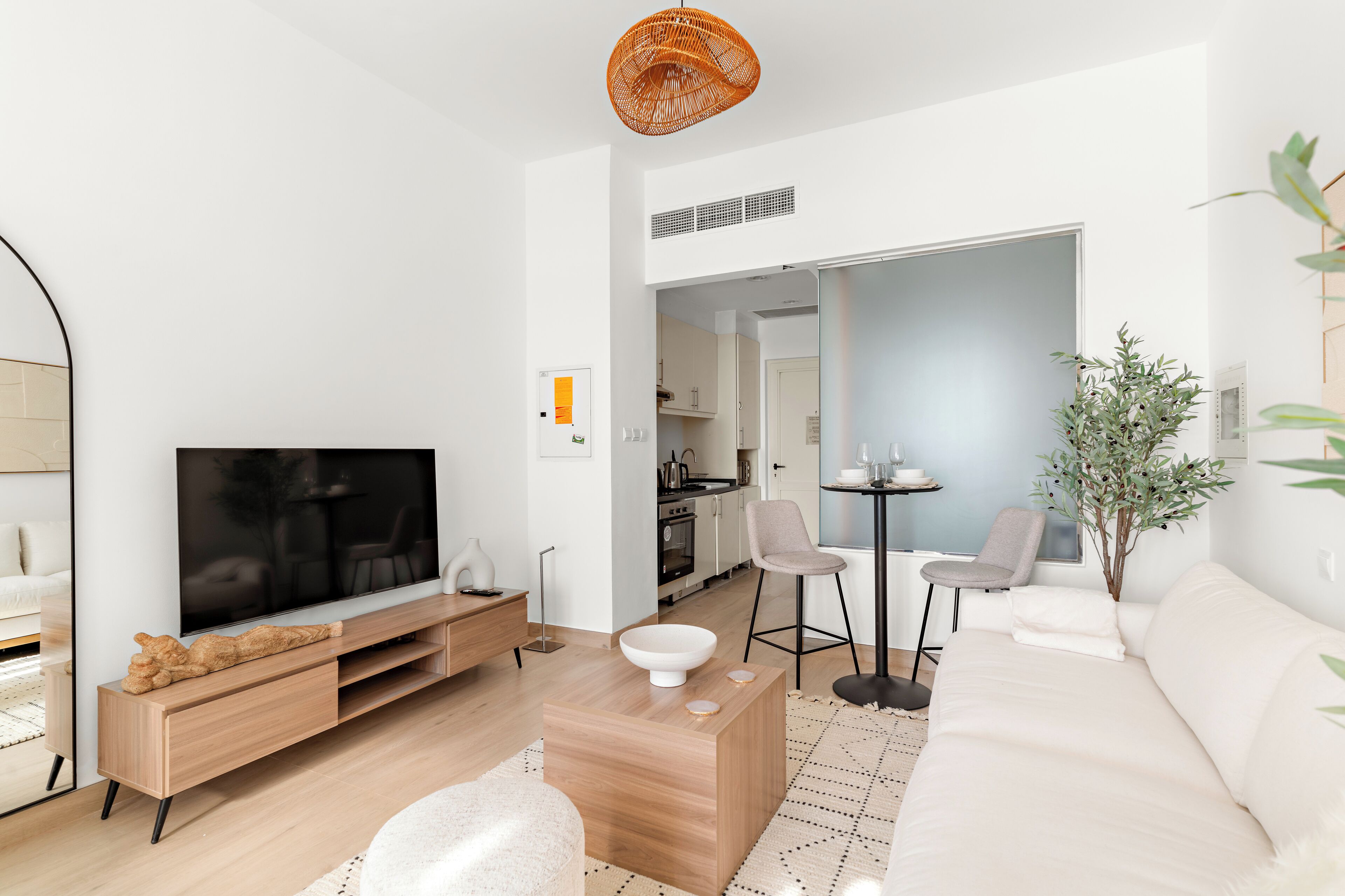 A modern living room with a flat-screen TV, wooden entertainment unit, white sofa, and a dining area with a small table and chairs.