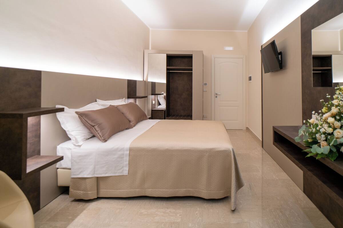 Habitación de hotel moderna con una cama grande, un armario, un televisor montado en la pared y un pequeño escritorio con una silla.