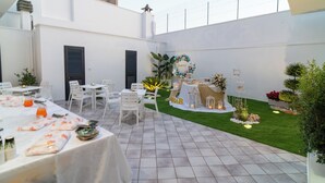 Une vaste terrasse extérieure avec des tables et des chaises blanches, un petit jardin agrémenté d’une arche décorative en forme de cœur, et un bâtiment blanc avec des volets en arrière-plan.