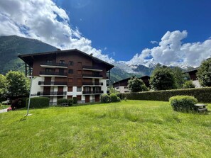 A multi-story building with balconies, surrounded by a well-maintained lawn and hedges, with a mountain backdrop.