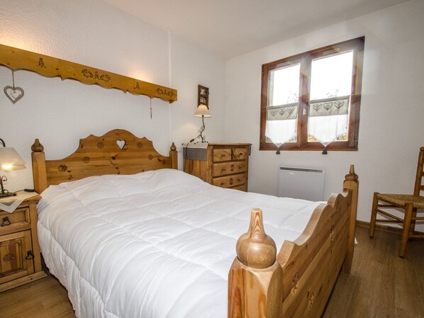 A wooden bed with a heart-shaped headboard, a wooden dresser, a nightstand with a lamp, and a window with a heart-patterned curtain.