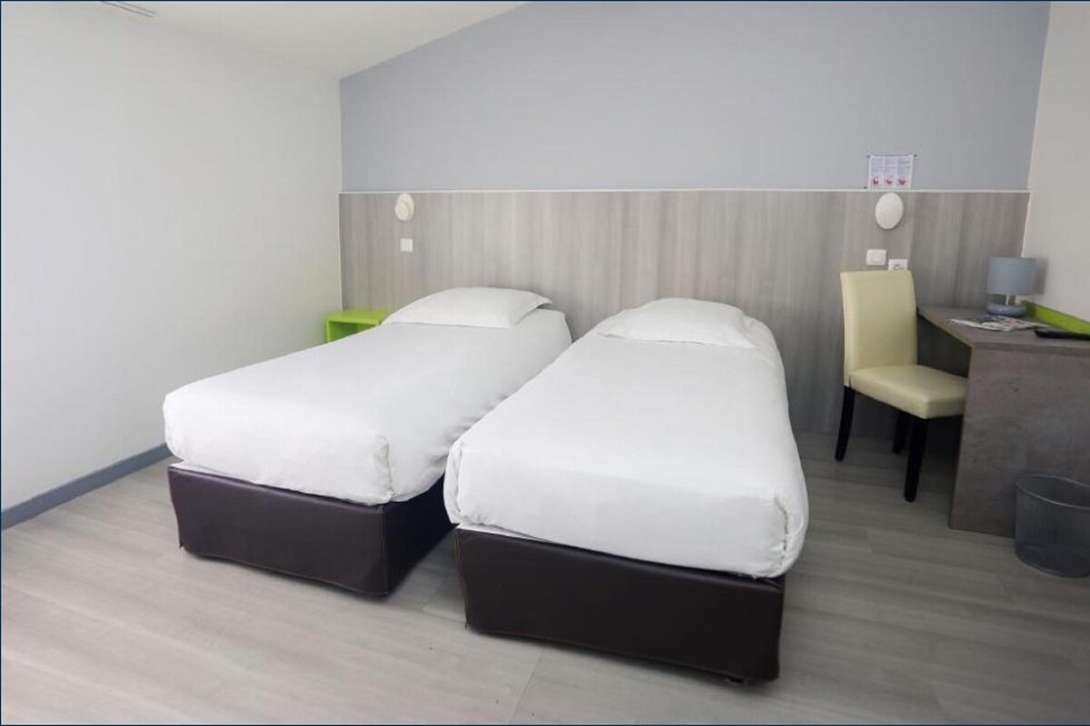 A hotel room with two single beds, a desk with a chair, a small trash bin, and a wall-mounted control panel.