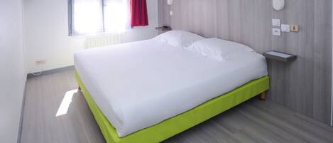 A neatly made bed with a green footboard in a room with light wooden flooring, a window with red curtains, and a wall-mounted TV.