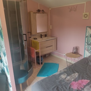 A bedroom with a bed, a small table with a lamp, a bathroom with a shower, and a mirror.