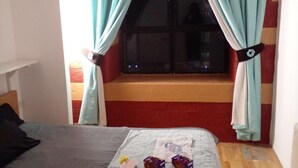 Ein Zimmer mit einem Bett, einem Vorhangfenster, einem kleinen Tisch mit Gegenständen und einer roten Wand.