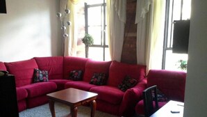 Ein Wohnzimmer mit einem roten Sofa, einem Holztisch, sowie einem kleinen Tisch mit einem Stuhl. Es gibt Fenster mit Vorhängen und eine Pflanze auf der Fensterbank.