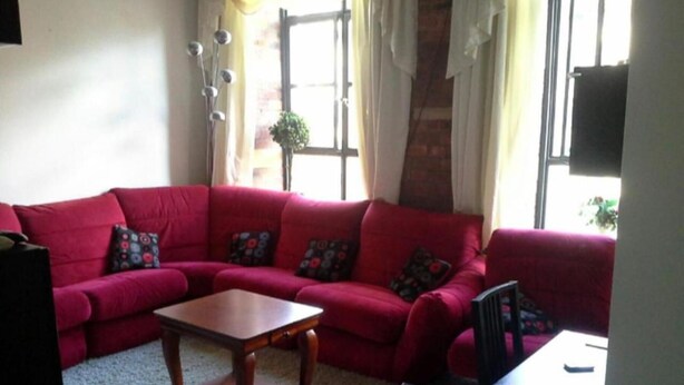 Ein Wohnzimmer mit einem roten Sofa, einem Holztisch, sowie einem kleinen Tisch mit einem Stuhl. Es gibt Fenster mit Vorhängen und eine Pflanze auf der Fensterbank.