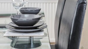 A dining table with a glass top, black plates, and a wine glass. The chair has a black leather seat. - Cosy 3 Bedroom Home With Wifi, Parking & Garden (Tilbury)