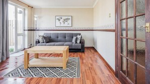 A living room with a grey sofa, a wooden coffee table, patterned rug, and a large window with curtains. - Cosy 3 Bedroom Home With Wifi, Parking & Garden (Tilbury)