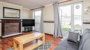 A living room with a wooden coffee table, a grey sofa, a television, and a large window with curtains. - Cosy 3 Bedroom Home With Wifi, Parking & Garden (Tilbury)