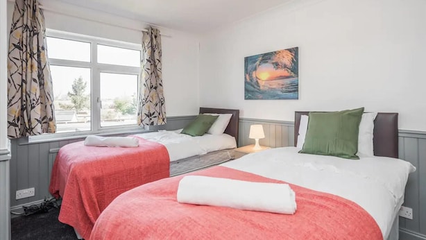 A hotel room with two beds, a window with curtains, a painting on the wall, and a lamp on the nightstand. - Cosy 3 Bedroom Home With Wifi, Parking & Garden (Tilbury)
