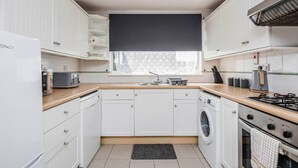 A compact kitchen with white cabinets, a built-in washing machine, a stove, and a window with a black blind. - Cosy 3 Bedroom Home With Wifi, Parking & Garden (Tilbury)