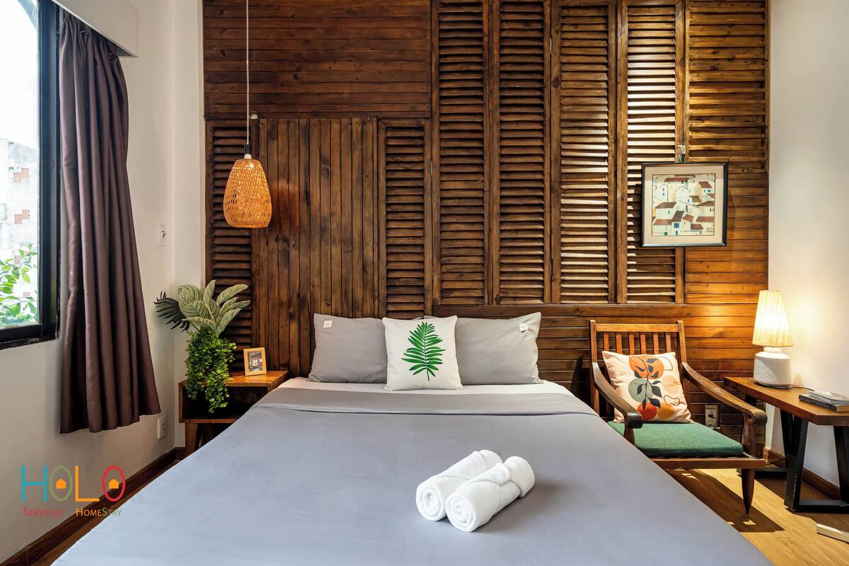 A hotel room with a bed, wooden headboard, a hanging lamp, a nightstand, a chair, a table, a lamp, a potted plant, and a framed picture on the wall.