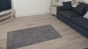 Una sala de estar con televisor de pantalla plana, un sofá gris oscuro, una mesita auxiliar con lámpara, un cuadro en la pared y una alfombra gris en el suelo.