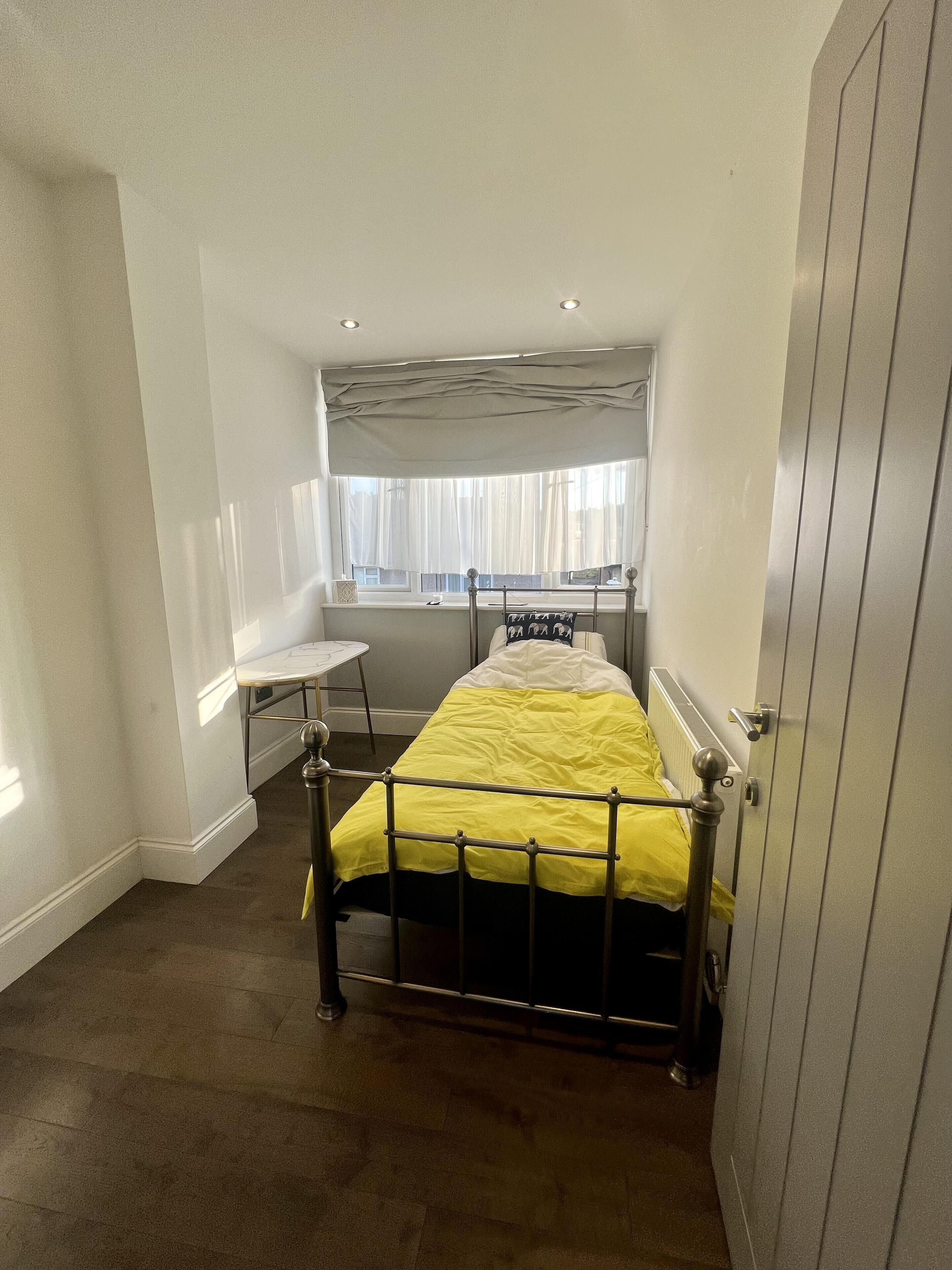 A small, narrow room with a single bed, a window with sheer curtains, a bedside table, and a radiator.