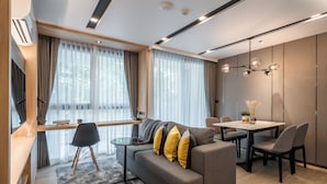 A modern living room with a sofa, dining table, and chairs. There is a large window with curtains, a ceiling light fixture, and a small table with a lamp.