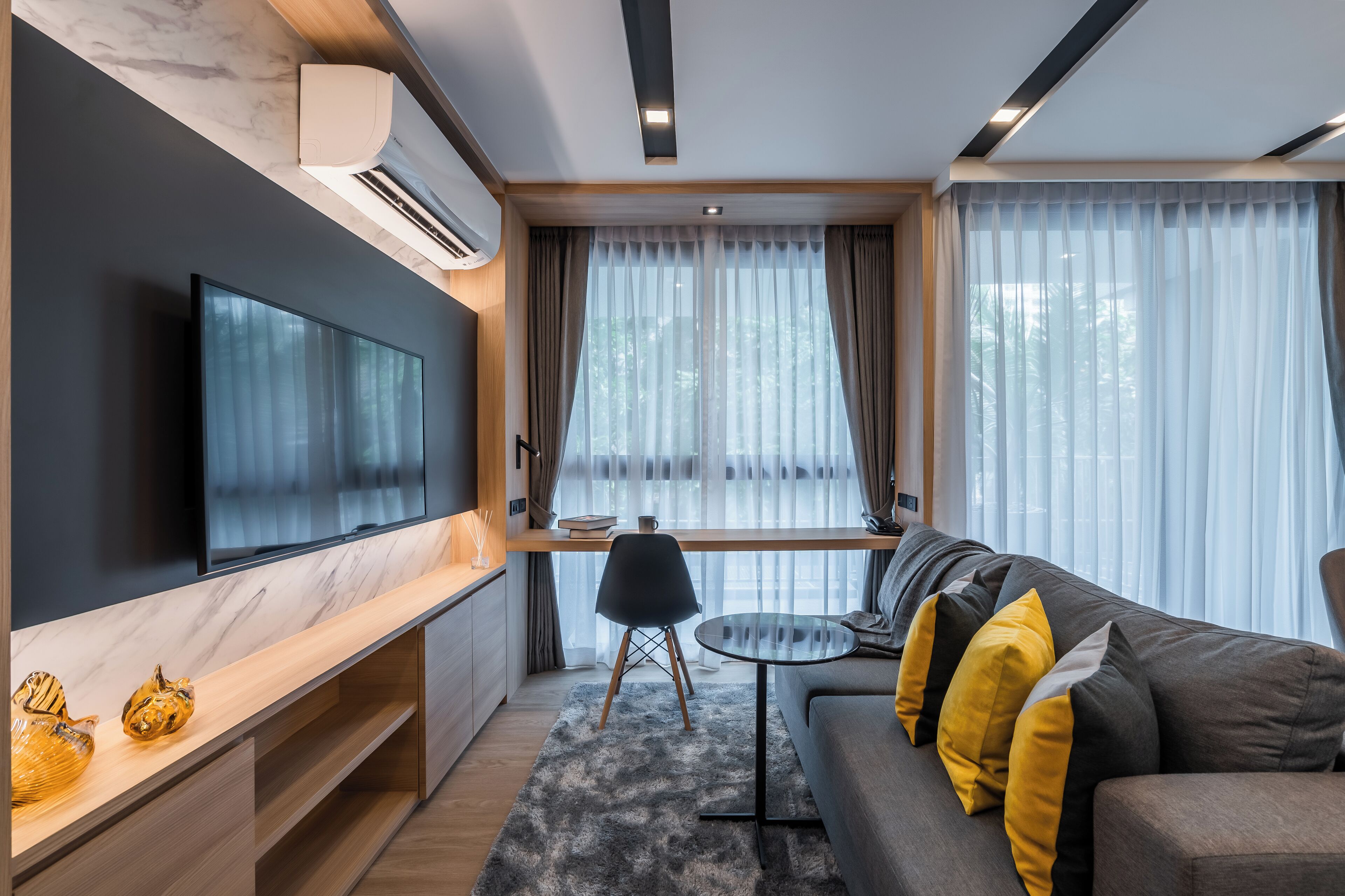 A modern living room with a flat-screen TV, a wooden desk, a sofa with yellow cushions, and large windows with sheer curtains.