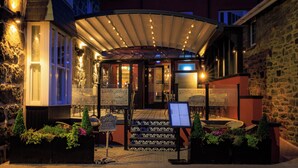 A covered outdoor seating area with a menu stand, stone walls, and a building entrance. - The Fanny Talbot  (Barmouth)