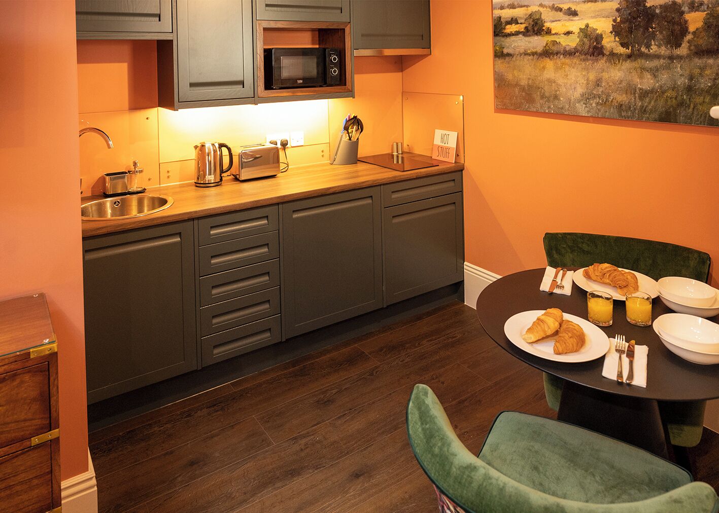 A compact kitchen with a microwave, toaster, and sink. There is a dining table with plates, glasses, and a croissant.