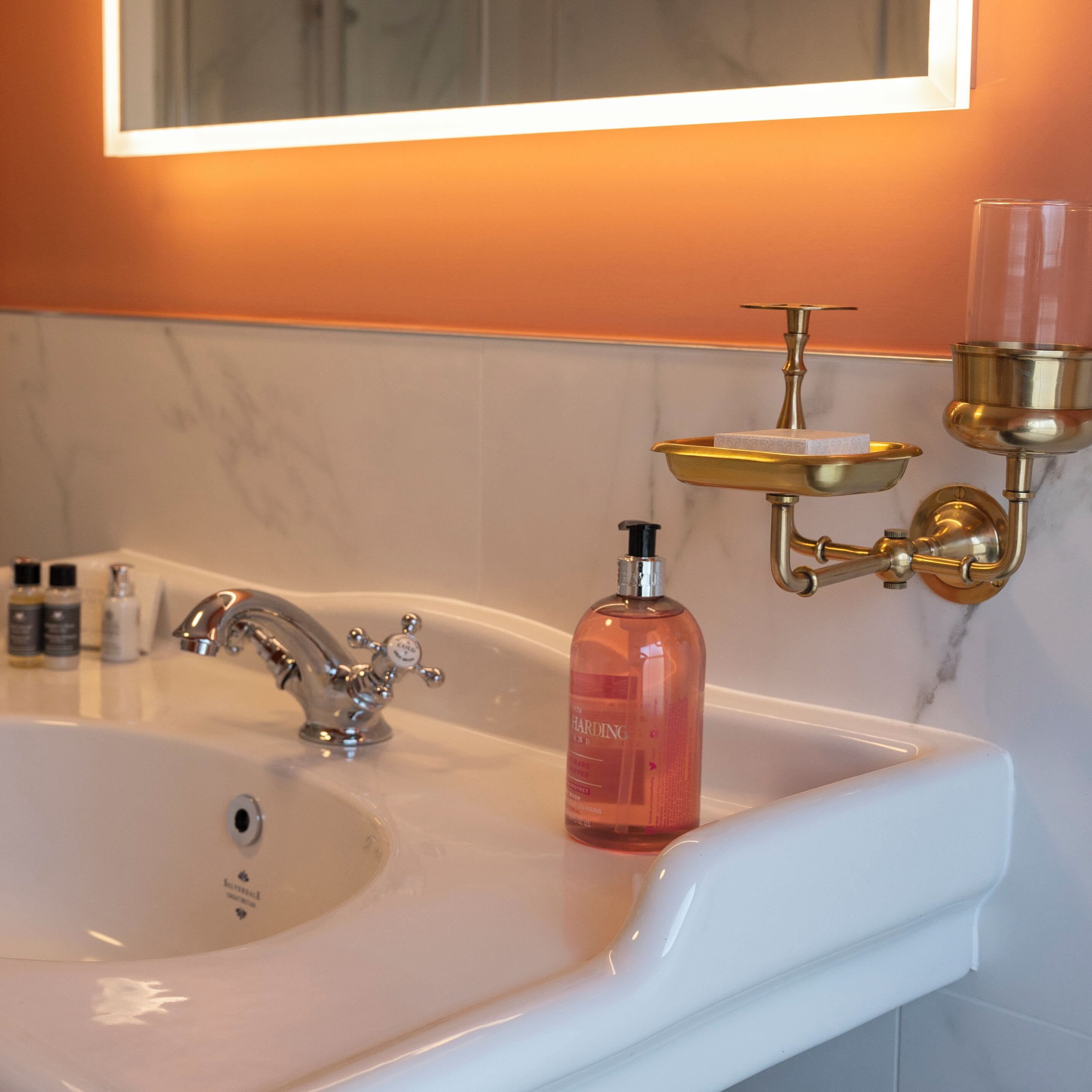 Un lavamanos de baño con un grifo moderno, un dispensador de jabón y un plato para jabón dorado fijado en la pared.