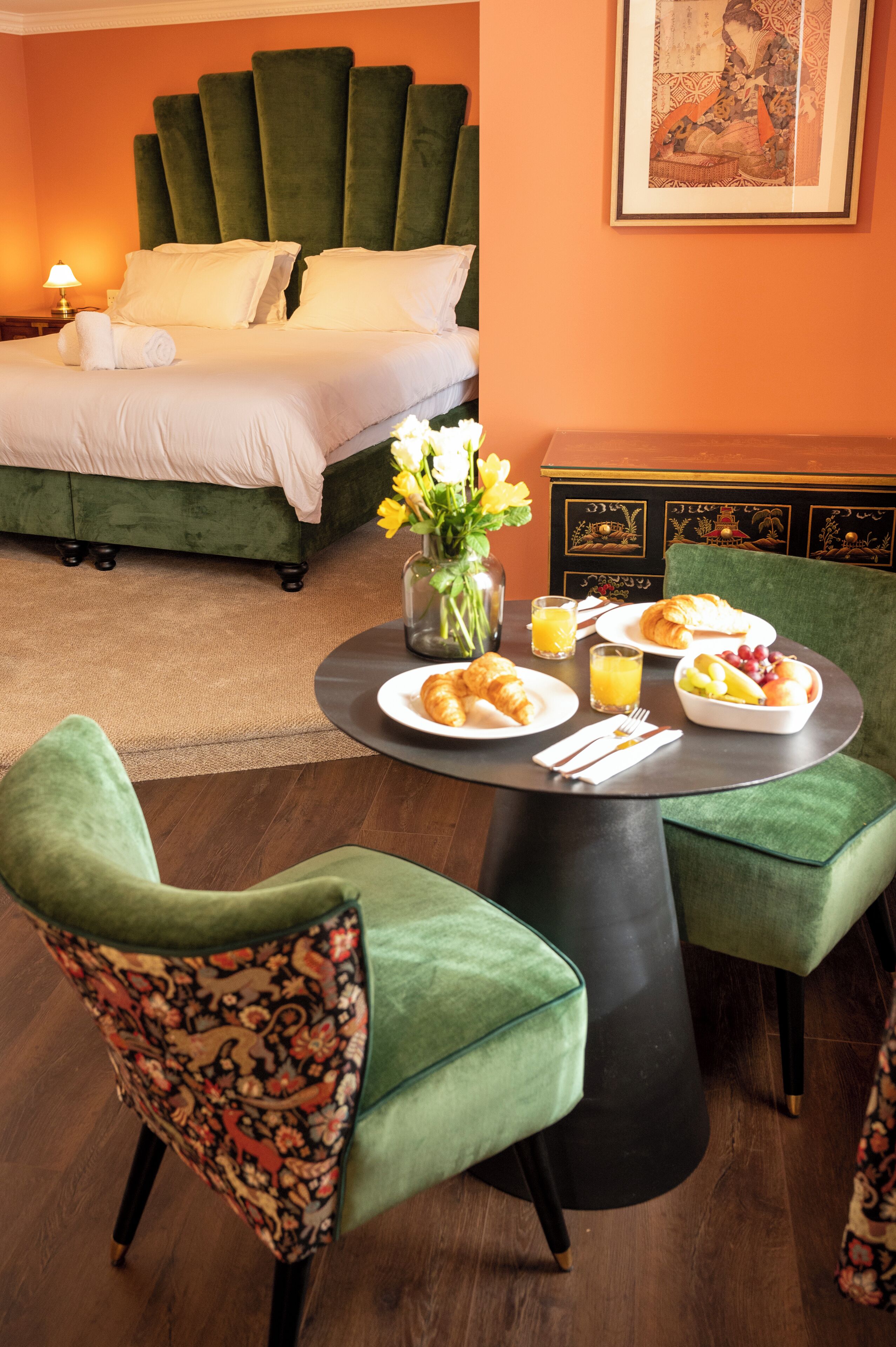 A hotel room with a bed, a dining table with breakfast, and green upholstered chairs.