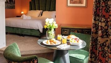 A hotel room with a bed, a dining table with breakfast, green upholstered chairs, and a painting on the wall.