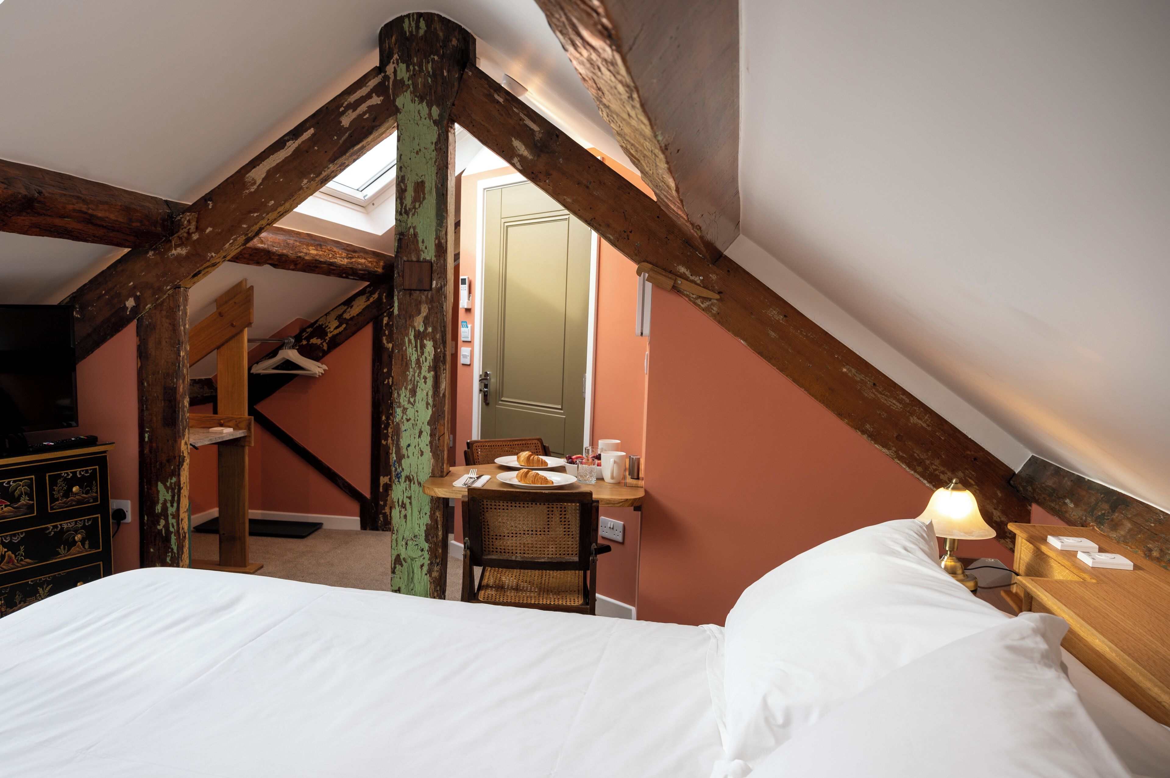 A bedroom with a sloped ceiling, wooden beams, a bed, a table with breakfast items, and a chair.