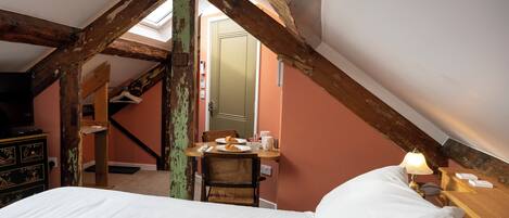 A bedroom with a sloped ceiling, wooden beams, a bed, a table with breakfast items, and a chair.